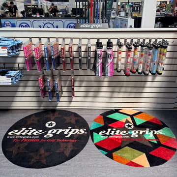Two circular Elite Grips floor mats displayed on a gray carpet inside a retail store. One mat is solid black with white text and a star logo, while the other features a colorful geometric pattern of red, green, yellow, and black triangles with the same branding.